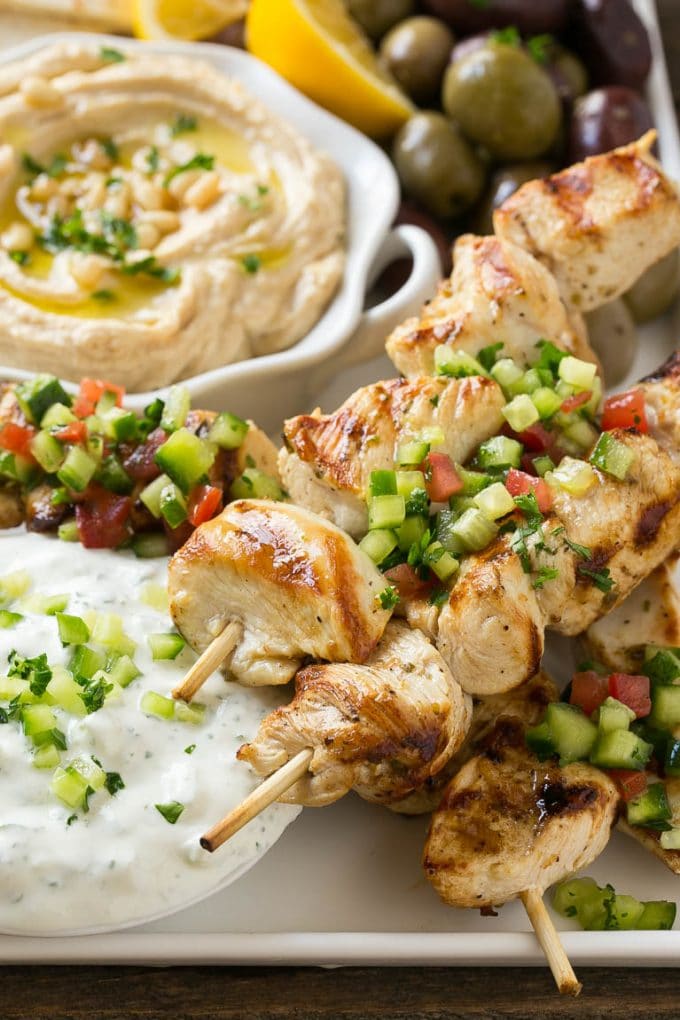 Greek Chicken Souvlaki Recipe | Easy Greek Chicken | Chicken Souvlaki | Greek Chicken Skewers | Easy Greek Chicken Skewers #chicken #kabobs #glutenfree #keto #lowcarb #grilling #bajap A tray of chicken souvlaki kabobs with olives, hummus, pita bread and yogurt sauce.
