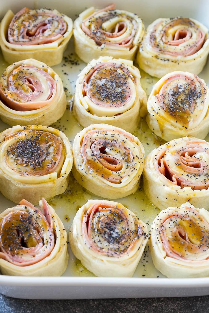 Ham and Cheese Pinwheels Recipe | Ham and Cheese Roll Ups | Ham and Cheese Rolls | Ham and Cheese Bread Rolls #ham #cheese #appetizer #pinwheels #bajap Dough rolled up with ham and cheese and topped with a poppy seed butter in a ceramic dish.