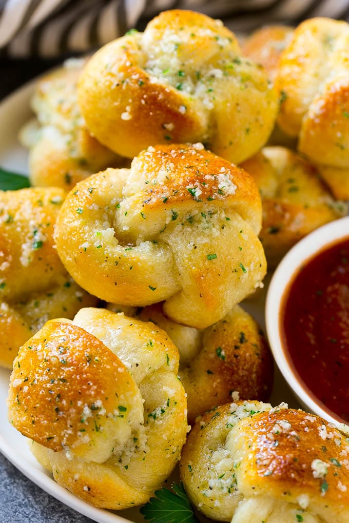 Garlic Knots Recipe | Garlic Rolls | Garlic Butter Rolls | Garlic Bread Recipe #garlic #garlicbread #garlicknots #breadsticks #bajap A plate of garlic knots slathered in butter next to a cup of marinara sauce.