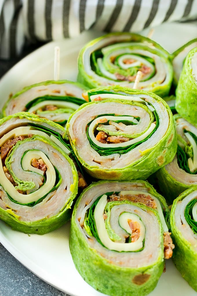 Turkey roll up sandwiches filled with cheese, avocado, bacon, turkey and spinach and stacked on a plate.