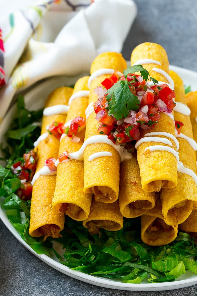 Chicken Taquitos Recipe | Baked Chicken Taquitos | Fried Chicken Taquitos | Mexican Appetizer | Chicken Tortillas #taquitos #chicken #appetizer #tortillas #mexicanfood #bajap A stack of chicken taquitos with a drizzle of sour cream and pico de gallo topping.