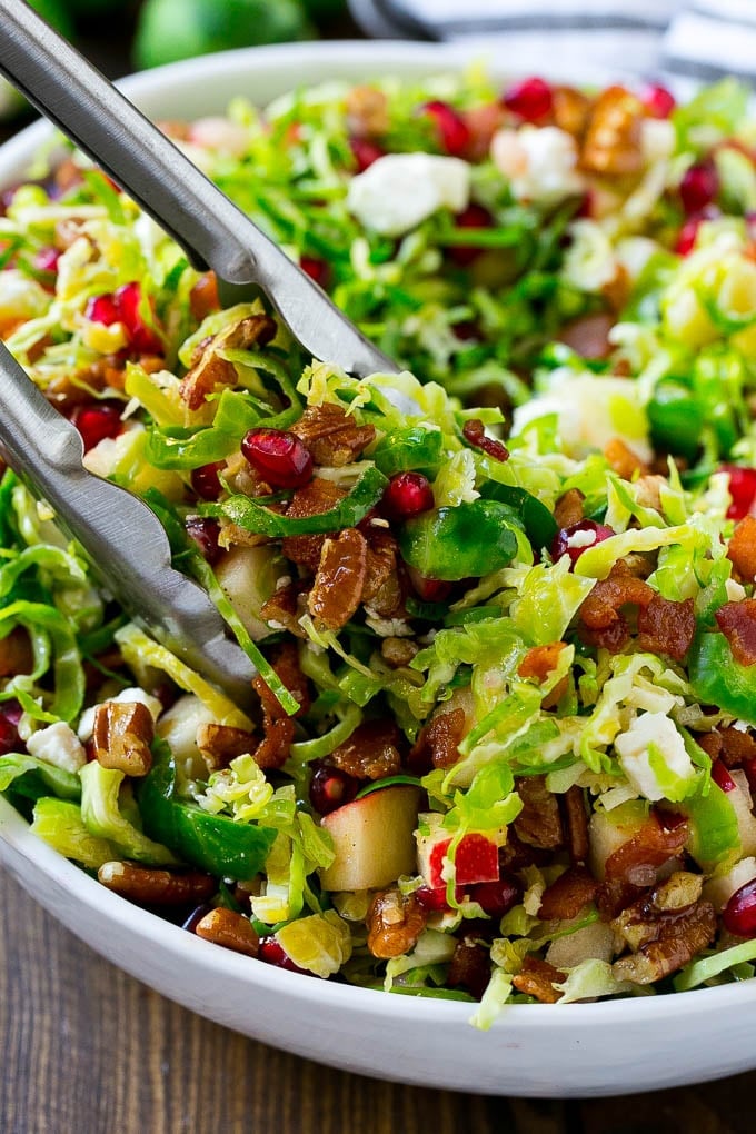 Brussels Sprout Salad Recipe | Shredded Brussels Sprouts Salad | Fall Salad #salad #brusselssprouts #bacon #thanksgiving #pomegranate #apple #dinner #bajap A bowl of brussels sprout salad with tongs picking some up out of the bowl.