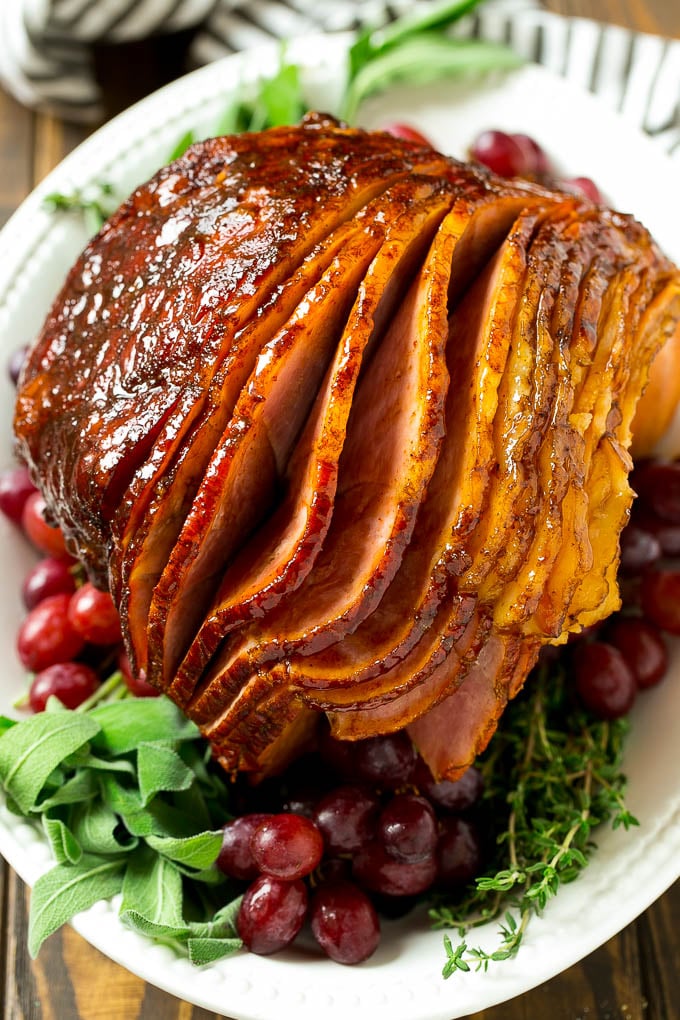 Brown Sugar Smoked Ham Recipe | Holiday Ham Recipe | Brown Sugar Glazed Ham #ham #christmas #thanksgiving #easter #smoker #dinner #brownsugar #bajap #glutenfree A brown sugar glazed smoked ham on a serving platter surrounded by grapes and fresh herbs.