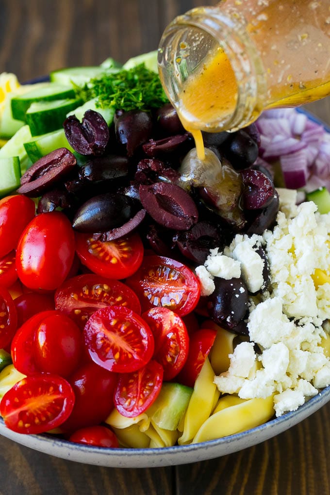 Greek Tortellini Salad Recipe | Pasta Salad | Tortellini Salad | Greek Salad #greek #tortellini #pasta #salad #cucumbers #olives #dinner #bajap Salad dressing being poured over a bowl of tortellini and vegetables.