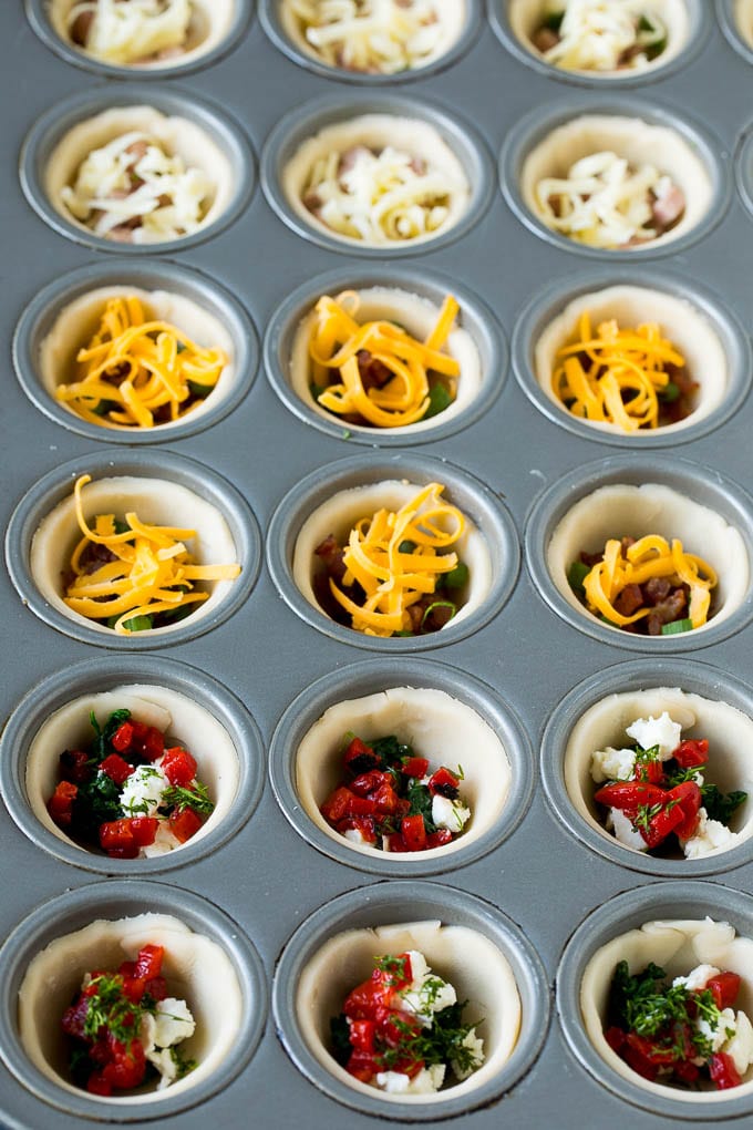 Mini Quiche Recipe #eggs #breakfast #brunch #appetizer #bajap Muffin tins filled with pie crust, vegetables, meat and shredded cheese.