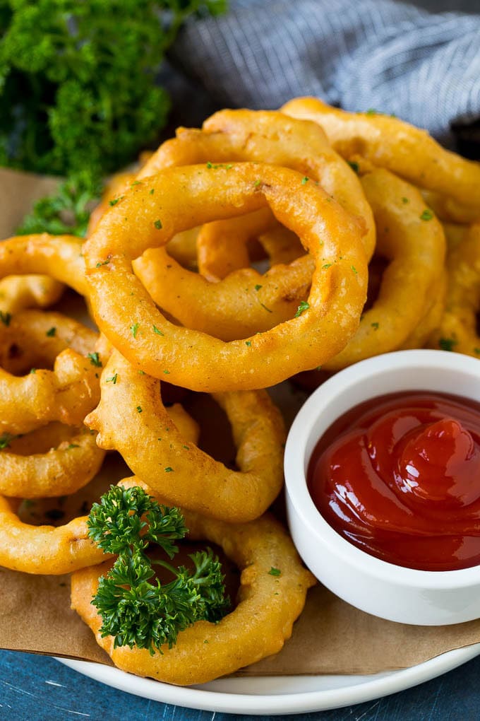 Crispy homemade onion rings that are fried in a flavorful beer batter. #onions #bajap #appetizer Fried onion rings on a plate with a side of ketchup.