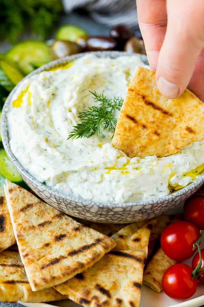 The best homemade tzatziki sauce with cucumber and herbs mixed into Greek yogurt. #appetizer #bajap A hand dipping pita bread into a bowl of tzatziki.