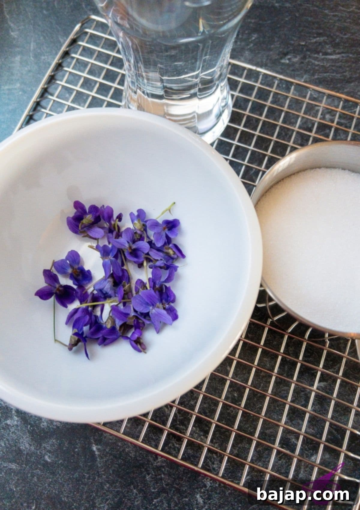 Fresh violets, superfine sugar, and water laid out as ingredients for candied violets.