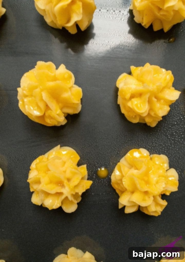 Close-up of Duchess Potato mounds being brushed with an egg yolk wash before baking, showing the detail of the piped shapes.