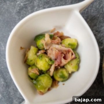 Brussels sprouts with fried bacon and chestnuts in a curved white bowl on a marble surface.