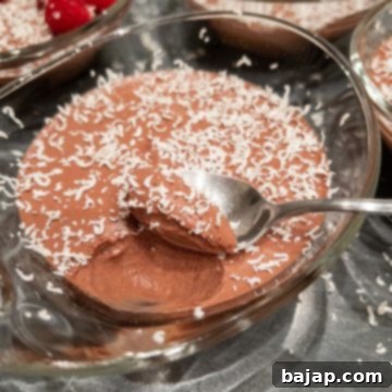 Small glass serving bowl filled with chocolate mousse and sprinkled with white chocolate shavings. Spoon scoops out of the mousse and reveals its perfect texture.