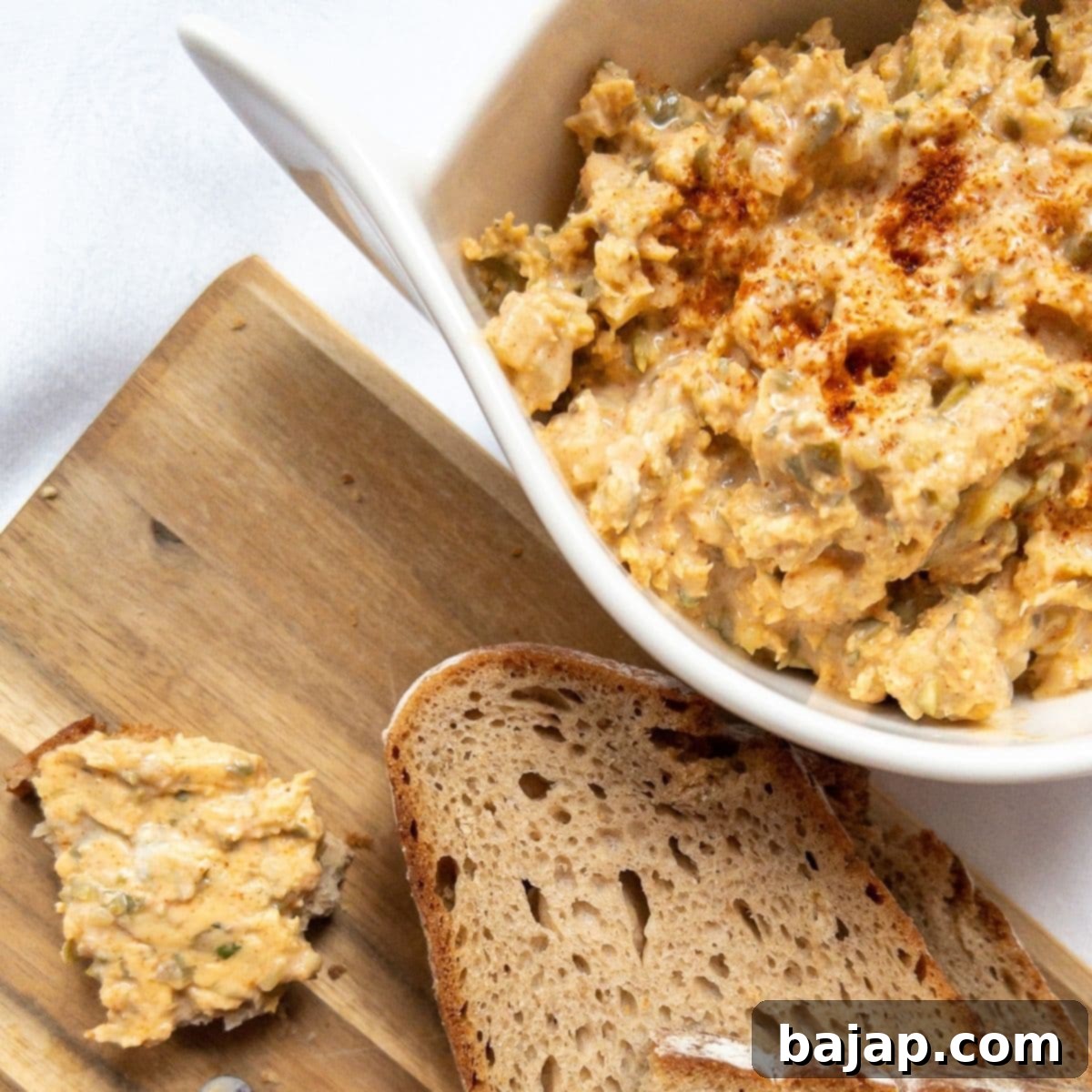 White bowl with Liptauer cheese spread on a white background next to a cutting board with bread slices.
