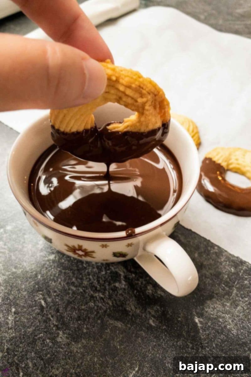 A hand dipping a cooled butter cookie into a bowl of melted dark chocolate couverture.