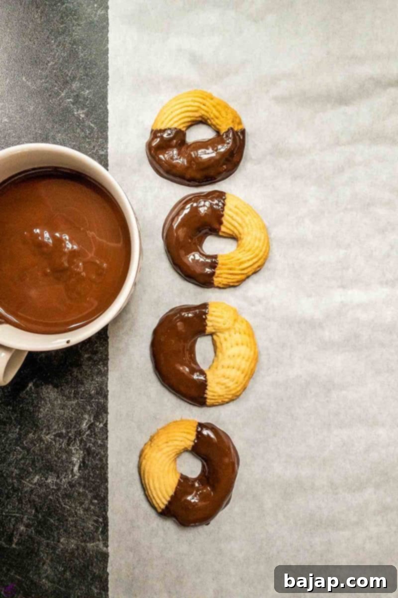A bowl of melted chocolate with four chocolate-dipped butter cookies placed on baking paper to dry, showcasing the glossy chocolate coating.