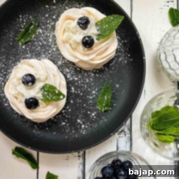 Berry Burst Pavlova Clouds 11 Meringue nests filled with whipped cream, decorated with blueberries and mint leaves, and dusted with powdered sugar. Placed on black serving plate.