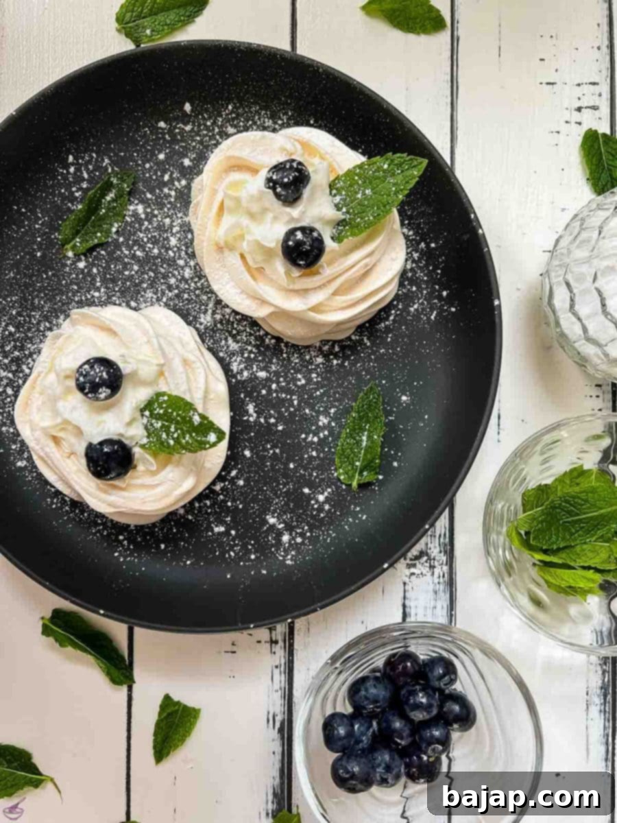 Berry Burst Pavlova Clouds 10 A beautifully arranged mini pavlova on a black serving plate, showcasing its fluffy whipped cream filling, vibrant blueberries, and delicate mint leaf, lightly dusted with powdered sugar, creating an irresistible dessert.