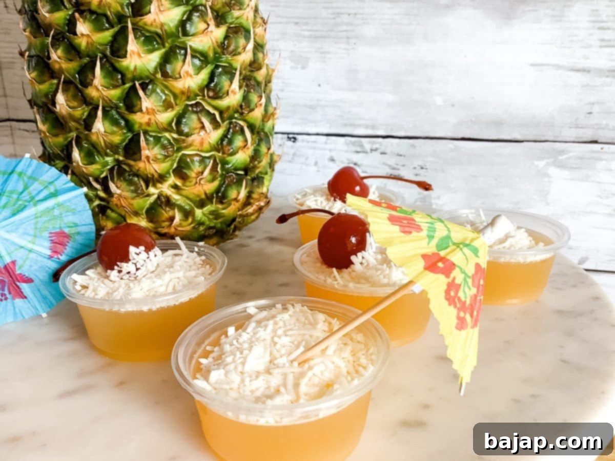Piña Colada Jello shots in small plastic cups, garnished with pineapple chunks and cherries.