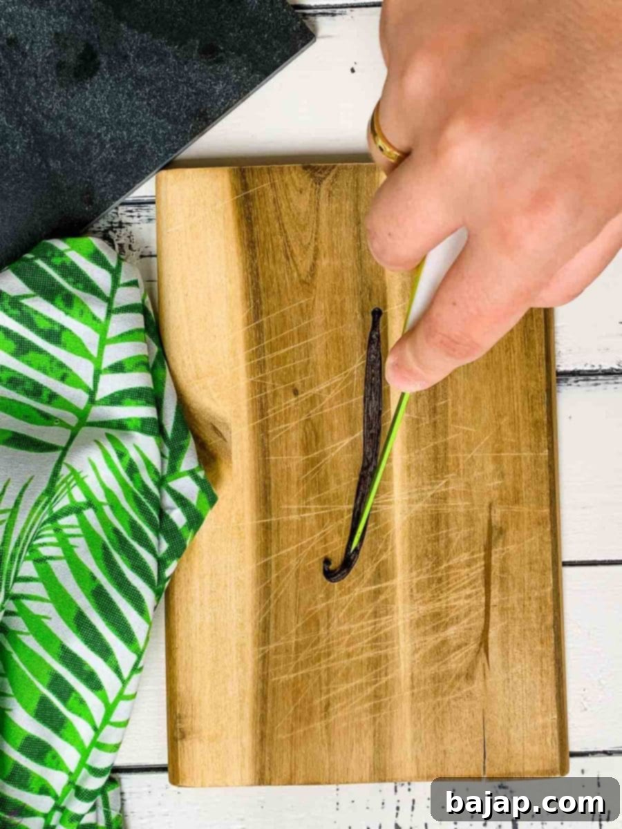 A sharp knife carefully cutting a vanilla bean open lengthwise on a wooden cutting board.