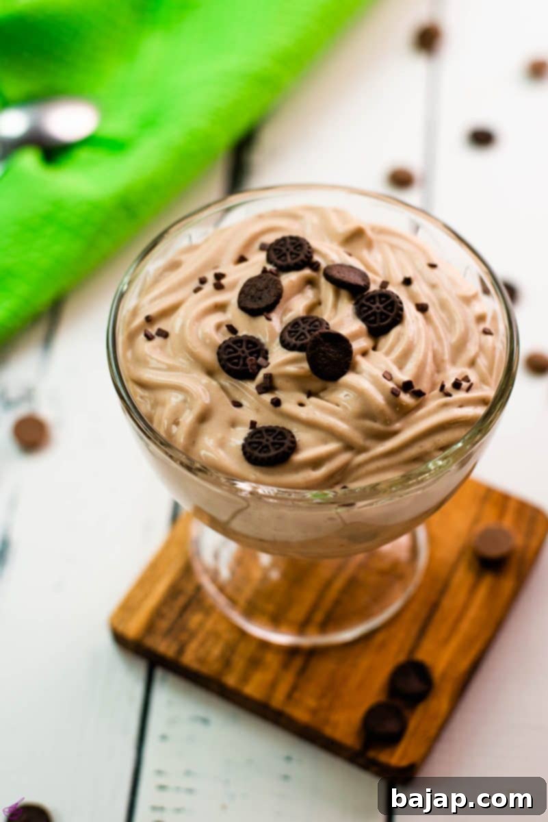 Beautifully textured chocolate cornstarch pudding in a dessert glass. Placed on a wooden coaster on a white wooden floor. Decorated with small pieces of biscuit.