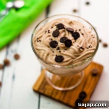 Beautifully textured chocolate cornstarch pudding in a dessert glass. Placed on a wooden coaster on a white wooden floor. Decorated with small pieces of biscuit.