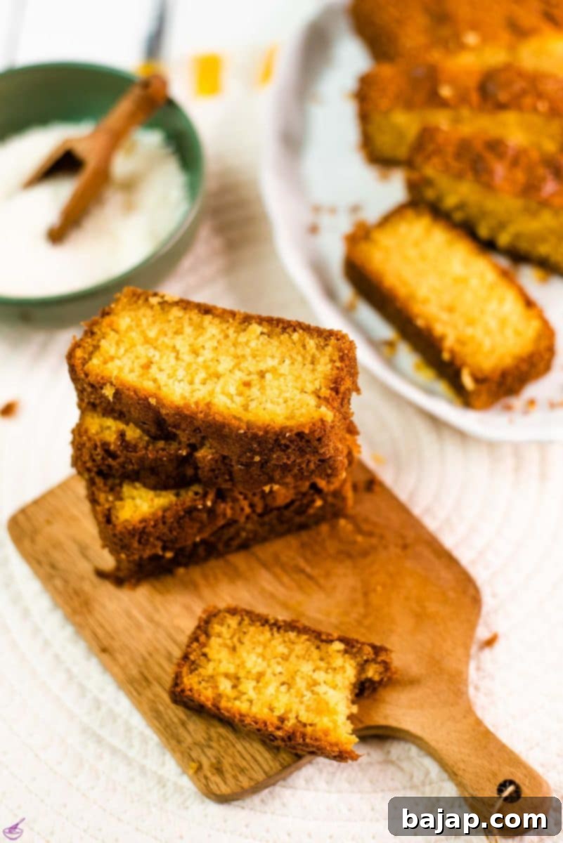 Beautifully stacked slices of moist coconut pound cake on an elegant wooden serving board. A bowl of shredded coconut and an entire sliced pound cake are blurred in the background, completing a tropical dessert scene.