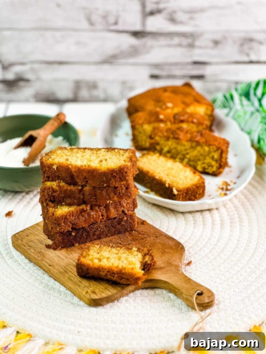 A captivating close-up of beautifully stacked slices of coconut pound cake on a wooden board. The rich, moist texture is evident, with shredded coconut visible throughout. In the blurred background, a bowl of coconut flakes and a whole sliced cake complete the inviting scene.