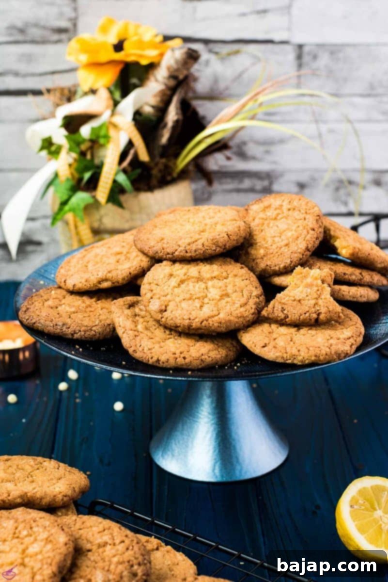 A pile of delicious lemon white chocolate chip cookies on a blue cake stand, next to fresh lemons and more cookies on a blue wooden board.