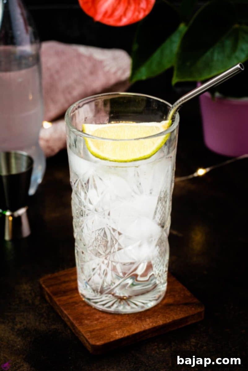 Highball glass filled with gin and tonic, garnished with lime, placed on wooden coaster.