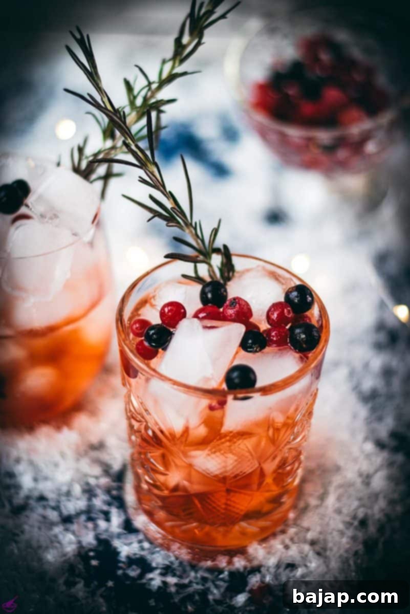 Two glasses filled with delicious cranberry Aperol spritz, placed on snowy ground, garnished with a delicious berry mix and rosemary.