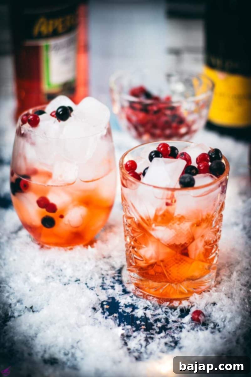 Two glasses filled with delicious cranberry Aperol spritz, placed on snowy ground, garnished with a delicious berry mix.