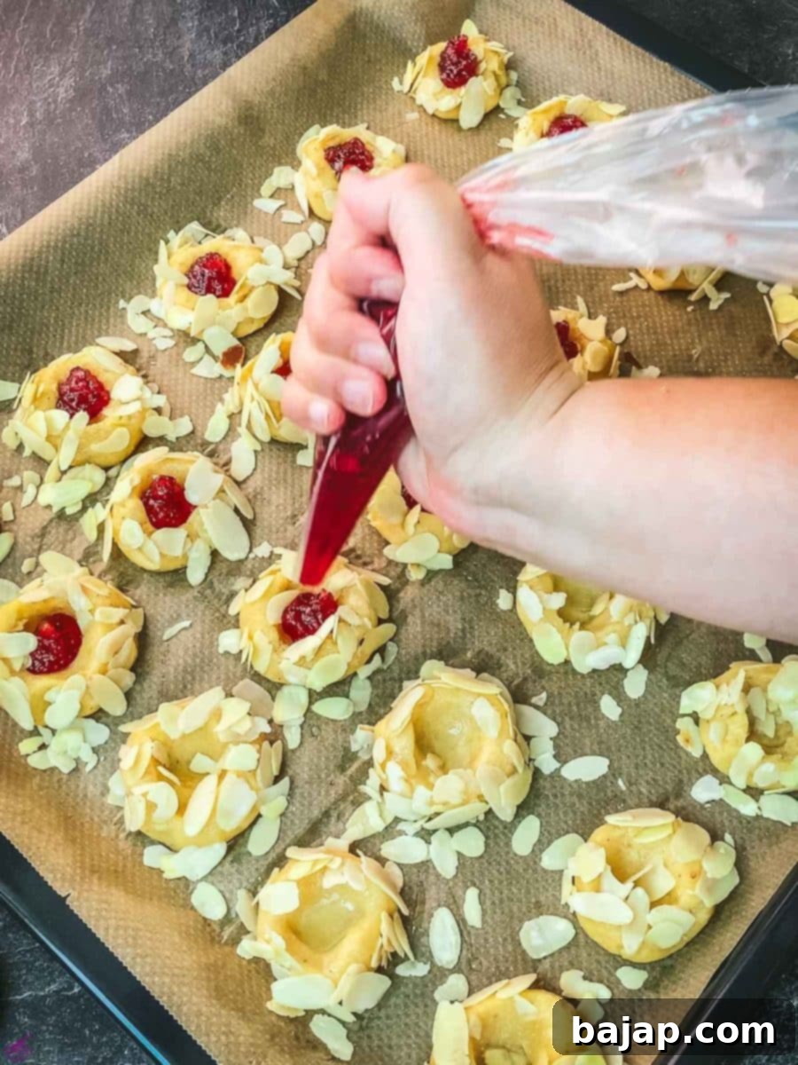Carefully filling the thumbprint indentations with vibrant raspberry jam, preparing the cookies for baking.