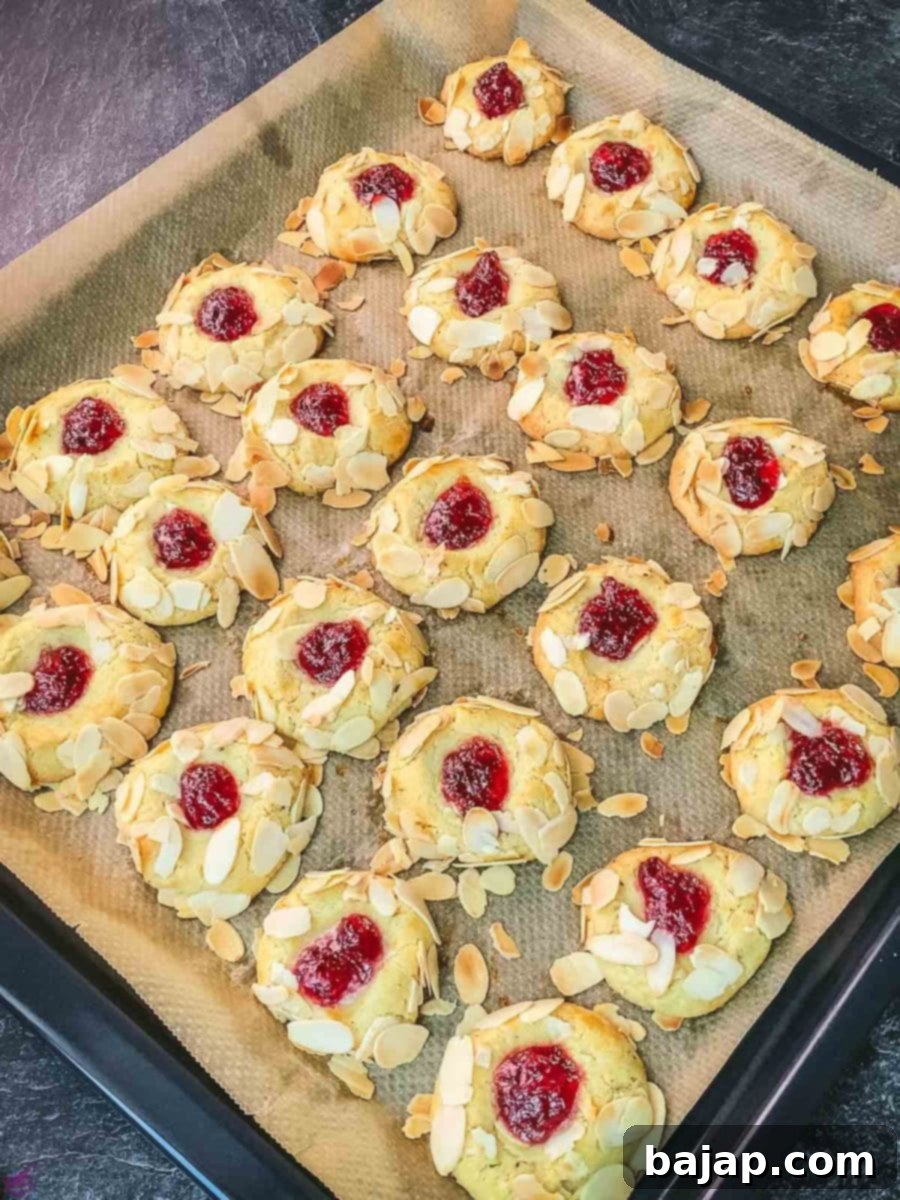 A baking sheet filled with golden-brown raspberry almond thumbprint cookies, fresh out of the oven, their jam centers glistening.