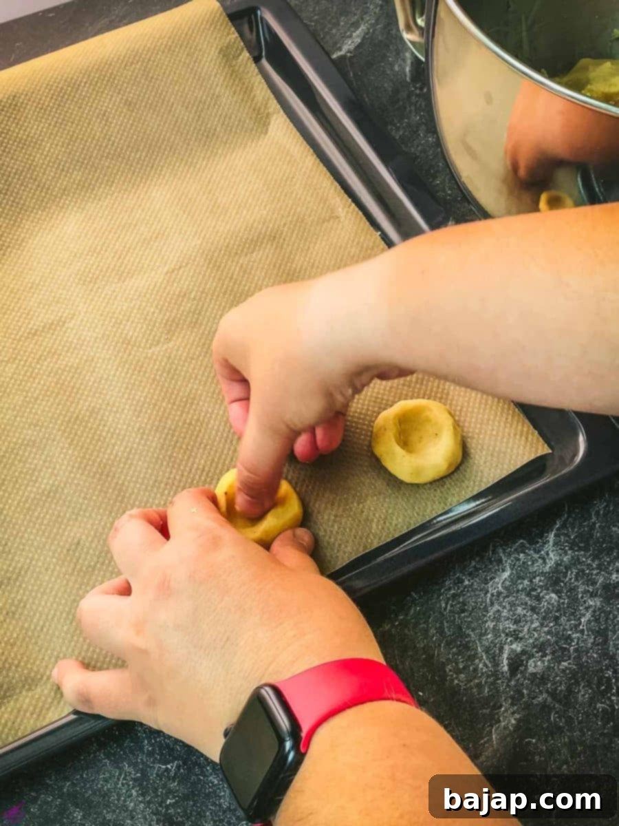 Pressing a thumbprint indentation into the center of each cookie ball on a baking sheet, preparing it for the jam filling.