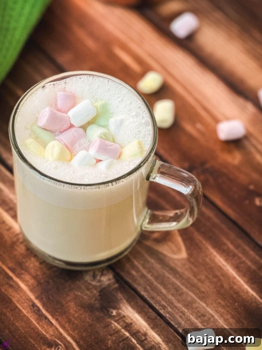 A clear glass cup brimming with rich, homemade white hot chocolate, elegantly topped with creamy frothed white chocolate and garnished with fluffy mini marshmallows. The perfect cozy drink.