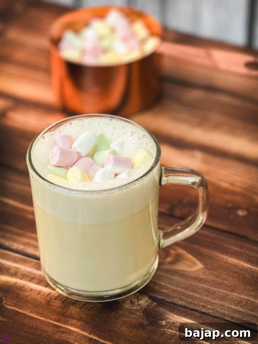 A beautifully presented glass cup of white hot chocolate, complete with a generous layer of frothed white chocolate and a sprinkle of mini marshmallows, inviting a warm and cozy sip.