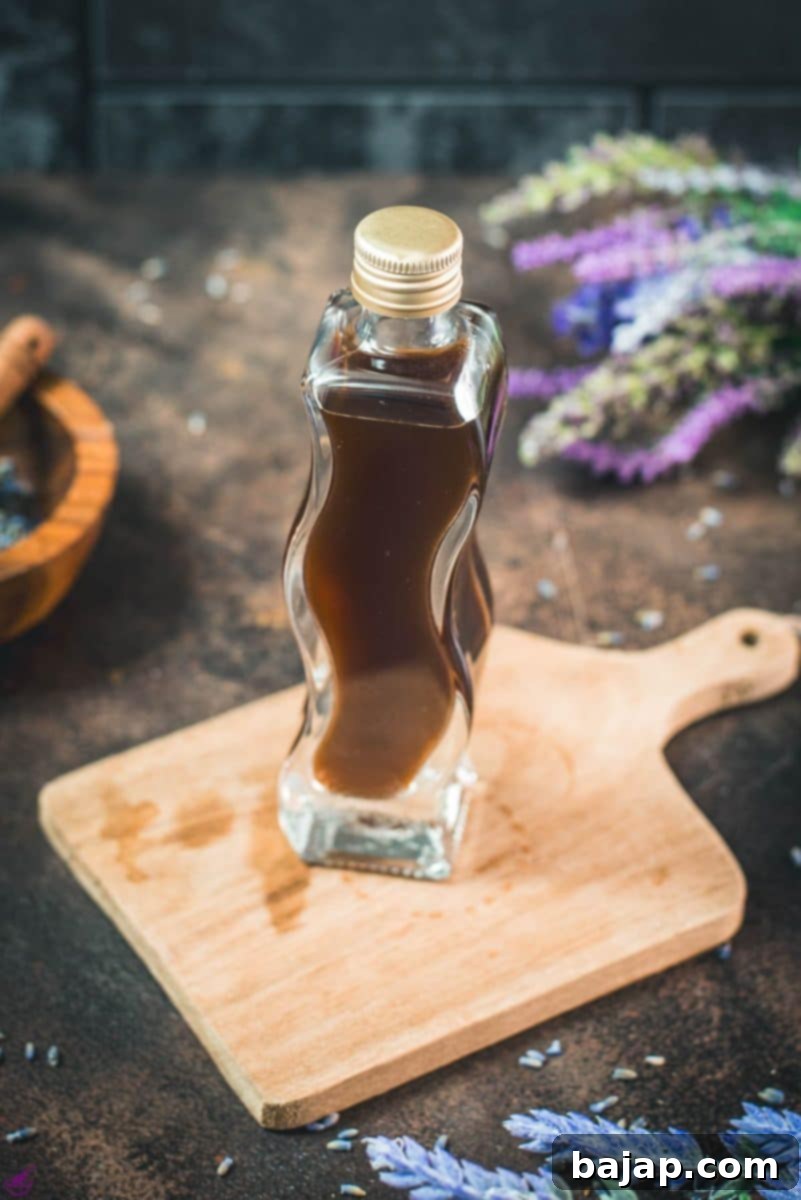 Curved bottle filled with homemade lavender extract on brown wooden board surrounded by lavender flowers.