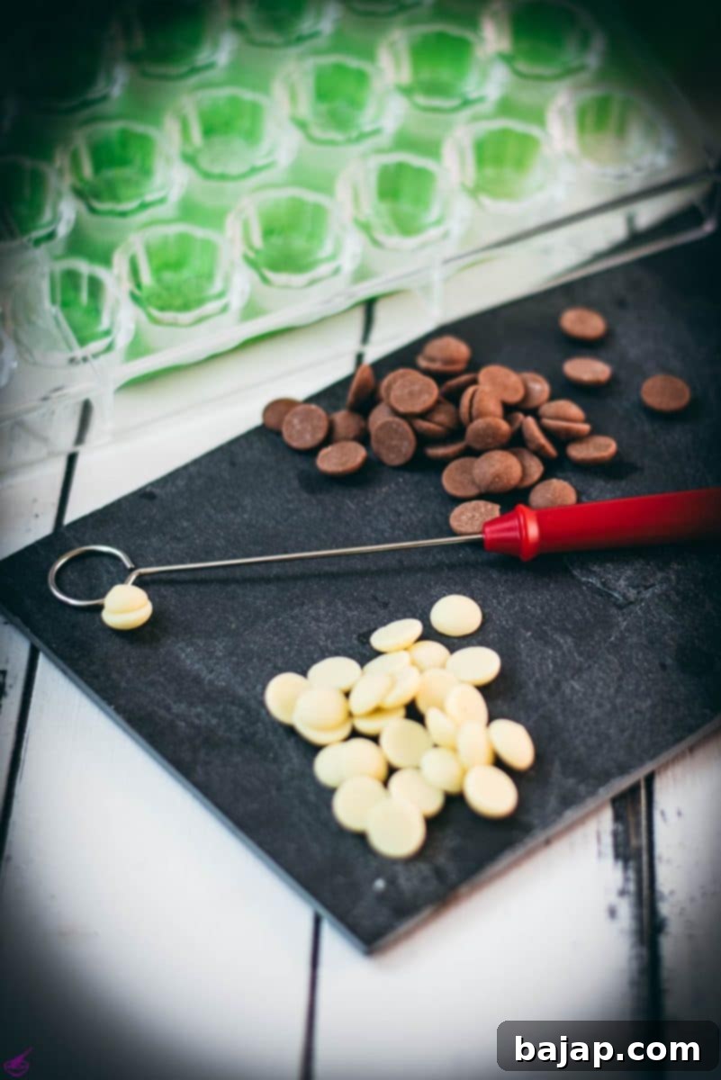 The Home Chocolatier's Essential Tool Kit 2 Black slate plate with chocolate dipper, molds, squeeze bottle and two kinds of chocolate couverture on white wooden ground.