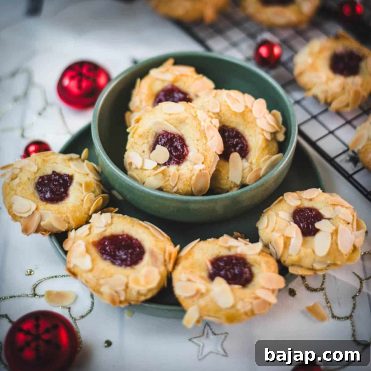 Delectable raspberry almond thumbprint cookies placed in a green bowl, next to red Christmas baubles.