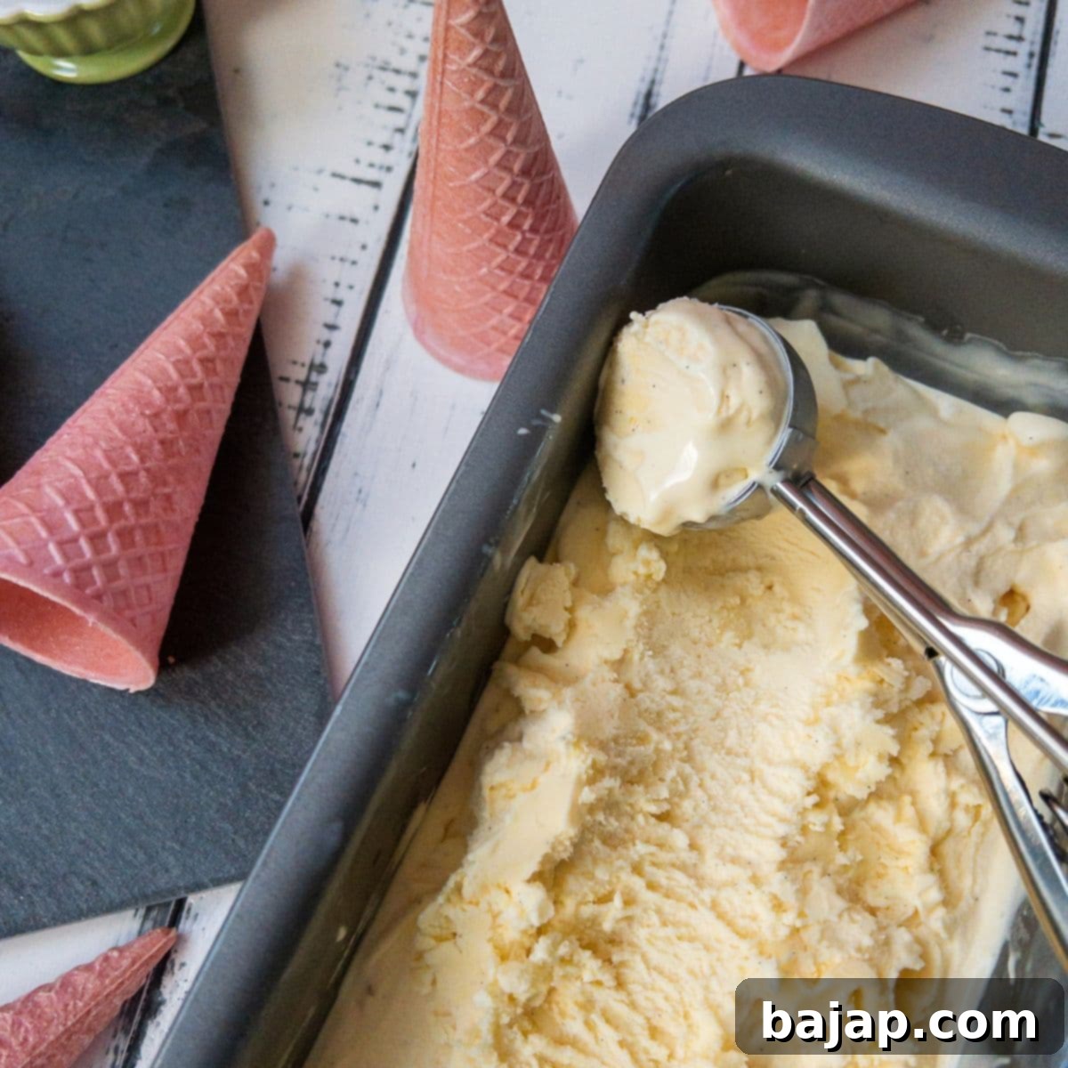 Loaf pan filled with homemade vanilla ice cream and scooped out vanilla icecream. With pink ice cream cones alongside.