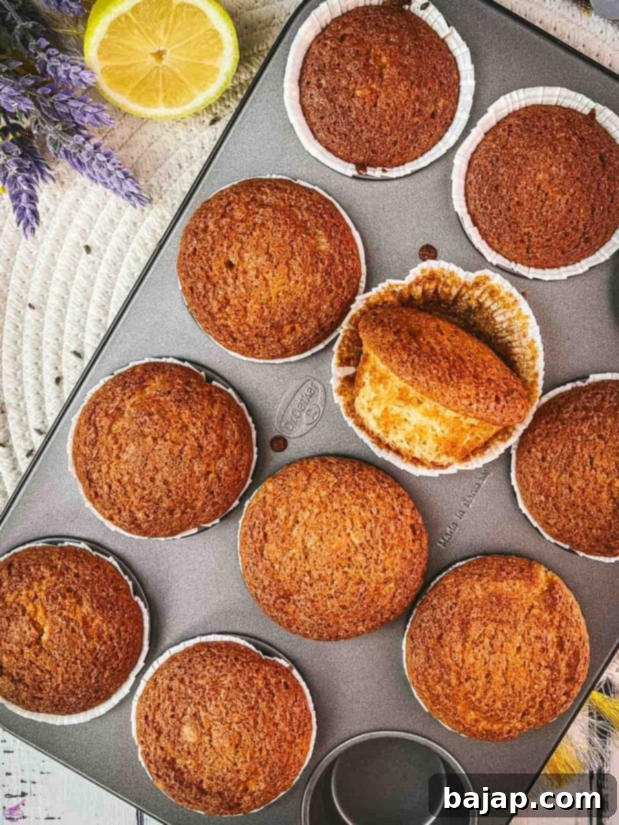 Freshly baked lavender lemon muffins cooling in a metal muffin rack, with one muffin playfully turned on its side to show its perfectly golden-brown exterior and delicate texture. These butter-free muffins are super fluffy and delicious, offering a subtle floral aroma with a zesty lemon tang.