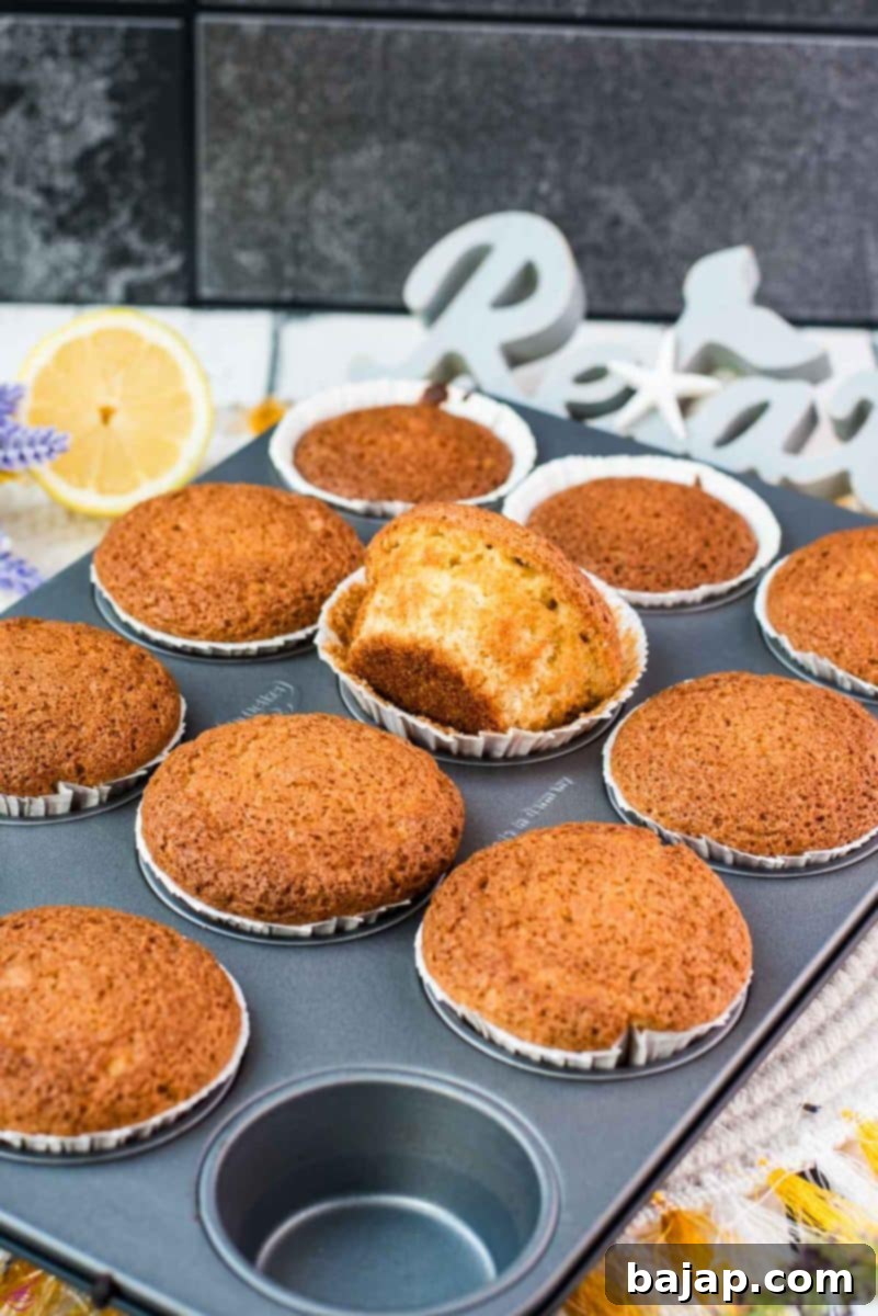 A close-up shot of several delightful lavender lemon muffins, still nestled in their metal muffin rack, with one muffin playfully turned to its side to reveal its moist interior. These butter-free, fluffy, and delicious muffins offer a perfect balance of subtle floral aroma and zesty lemon tang, making them an irresistible homemade treat.