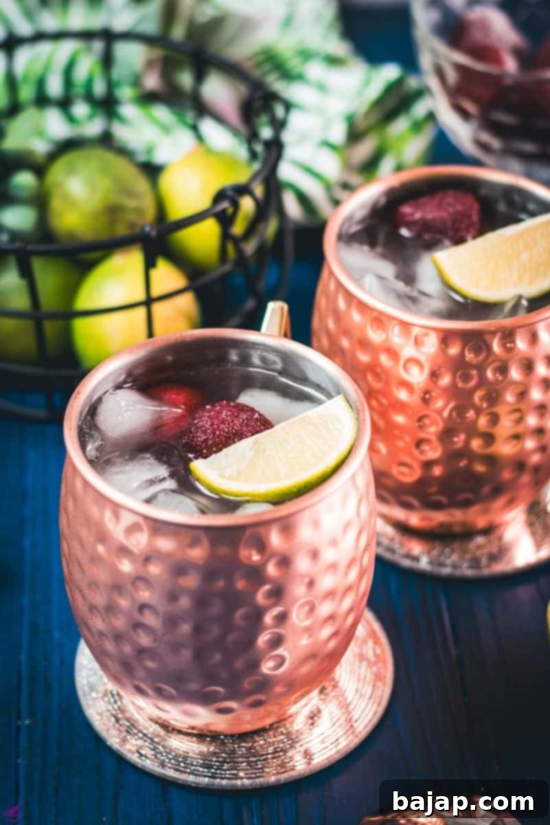 Gorgeous copper cups filled with delicious strawberry Moscow mule placed on blue wooden ground, garnished with fresh strawberries and lime wedges.