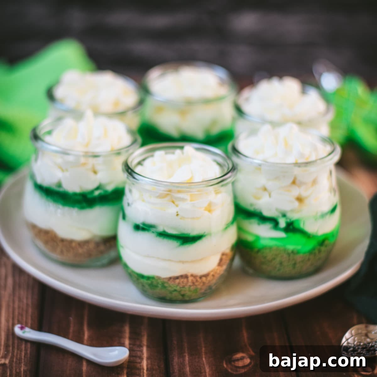 Gorgeous no bake lime cheesecakes in a jar for celebrating St. Patrick's Day! Placed on a white plate sitting on a brown wooden floor.