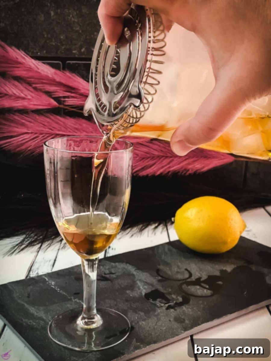 Slowly and carefully pouring the stirred Martinez cocktail through a strainer into a chilled Nick & Nora glass, ensuring a smooth, ice-free drink.