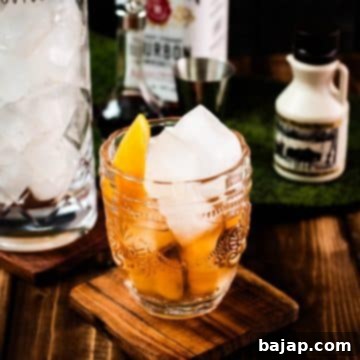 Golden Maple Old Fashioned 12 Gorgeous glass filled with maple old fashioned on wooden coaster. The drink is garnished with an orange zest and placed next to a stirring glass and a maple syrup bottle.