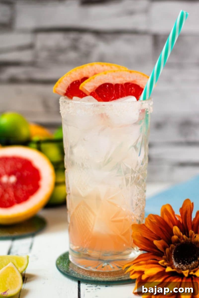A beautifully presented Paloma cocktail, garnished with fresh grapefruit slices and a straw, sitting on a mirrored coaster, perfect for a sunny day.