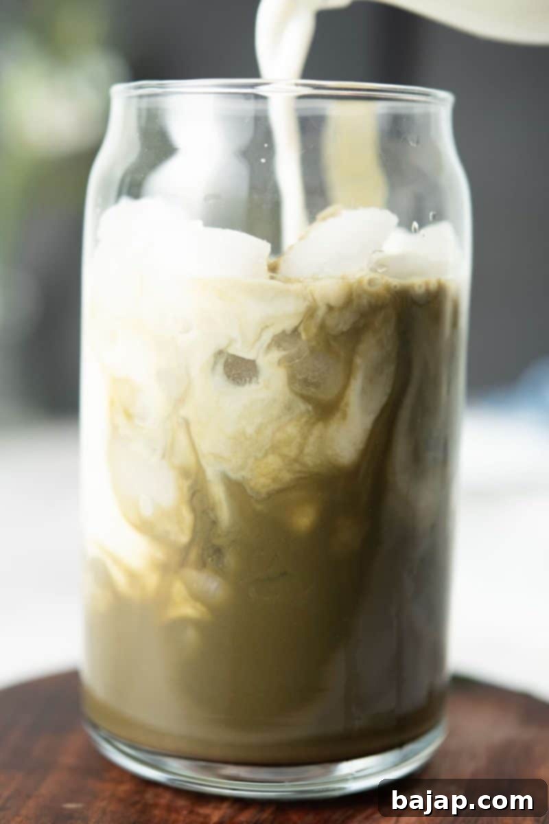 Pouring frothy oat milk into a glass with a vibrant green matcha mixture.
