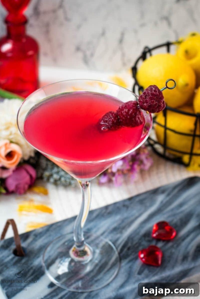 A close-up shot of a raspberry lemon drop martini, showcasing its vibrant pink hue, garnished with a single cocktail spike holding three fresh raspberries, set against a blurred background.