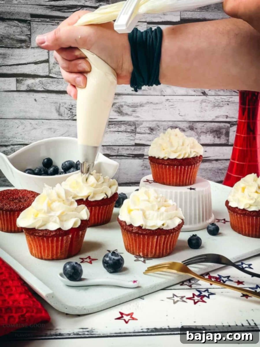 Hand piping white crème fraîche cream on top of the red muffin sponge, using a piping bag with a star tip, creating a beautiful swirl.