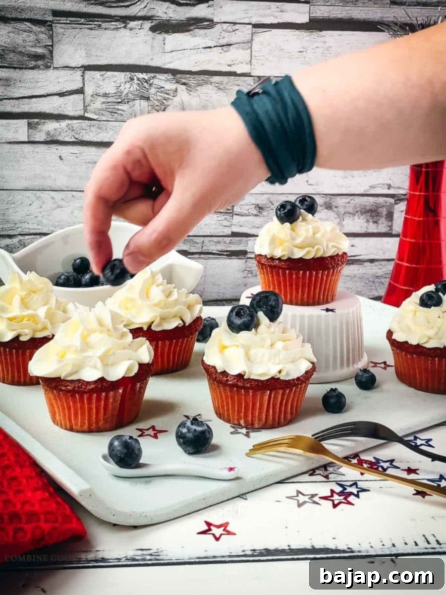 Hand placing a single fresh blueberry on top of the white cupcake frosting, adding the final patriotic touch.